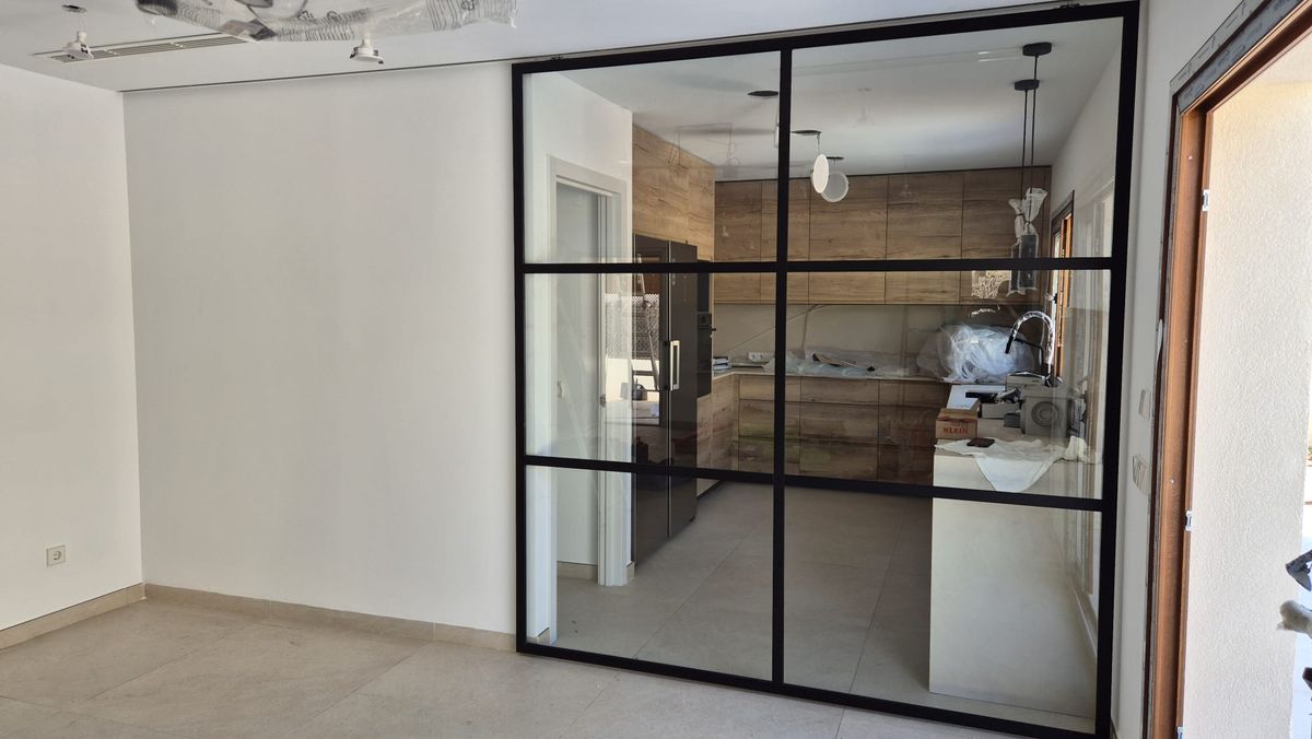 Cocina reformada por Las Marquesinas: mobiliario de madera, encimera clara y cerramiento acristalado de perfil negro que la separa del salón